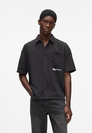 Ung mand iført en struktureret sort kortærmet skjorte med knapper og brystlomme samt "Karl Lagerfeld Jeans"-logo, kombineret med mørke jeans.