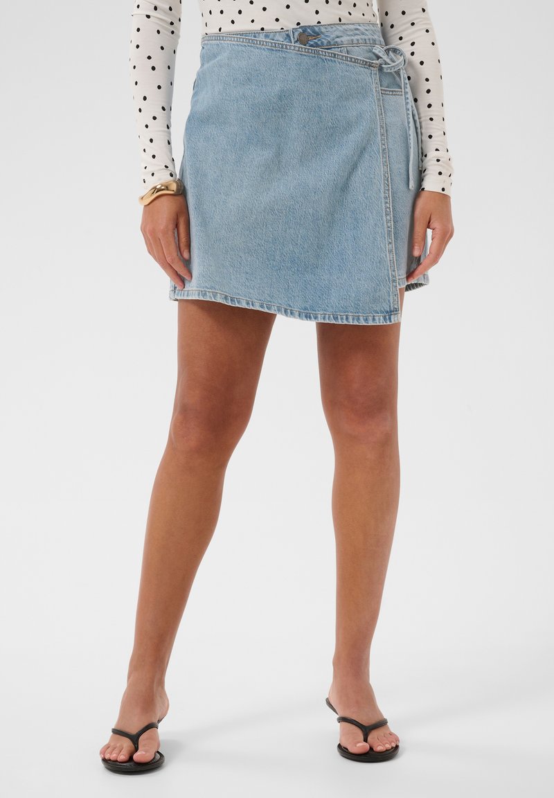 Femme portant une jupe portefeuille en jean bleu clair, un haut à manches longues à pois, un bracelet en or et des sandales noires en forme de tongs, debout devant un fond blanc.