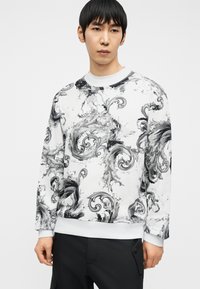 Άνδρας που φοράει λευκό φούτερ με μαύρα και γκρι ελικοειδή μάρμαρα και μοτίβα σε στιλ μπαρόκ, συνδυασμένο με μαύρο παντελόνι.