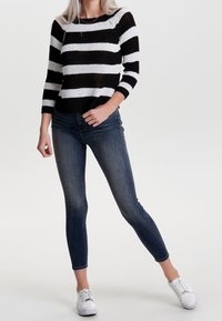 Femme portant un pull à rayures noires et blanches, un jean skinny bleu foncé et des baskets blanches, debout avec une jambe pliée.