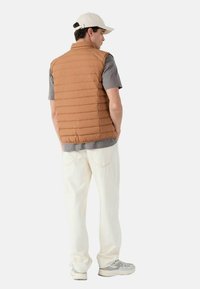 Gilet trapuntato color tan sopra a una maglietta grigia a maniche corte, abbinato a pantaloni bianchi e sneakers grigio chiaro. Presenta un colletto alla coreana e tessuto strutturato.