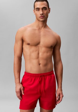 Homme musclé debout devant un fond gris, portant un short de bain rouge vif avec une taille à cordon, regardant droit devant lui.