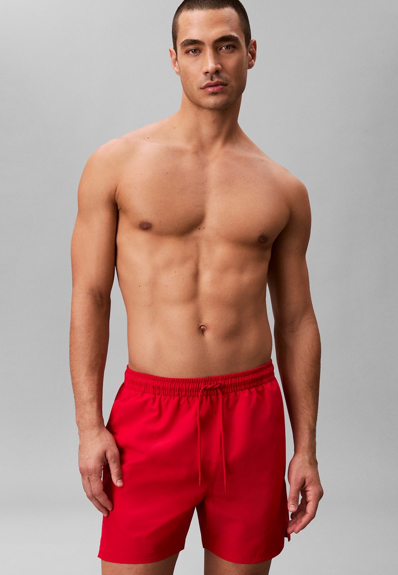 Homme musclé debout devant un fond gris, portant un short de bain rouge vif avec une taille à cordon, regardant droit devant lui.