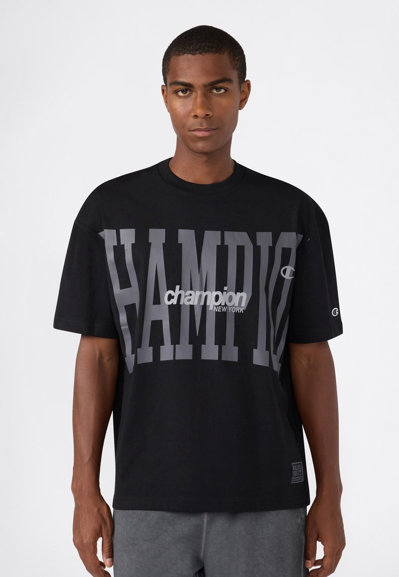 Giovane uomo che indossa una t-shirt oversized nera della Champion con grande testo del marchio grigio e bianco, in piedi davanti a uno sfondo bianco semplice.