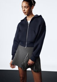 Marineblå kort zip-up hettegenser med stor hette og frontlomma, kombinert med grå striper skjørt med plisserte detaljer.