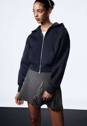 Navy cropped zip-up hoodie with a large hood and front pocket, paired with a grey pinstripe skirt featuring pleated accents.