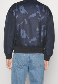 En person som bär en marinblå bomberjacka med ett nätliknande kamouflagemönster på ryggen, tillsammans med ljusblå denimjeans, sedd bakifrån.