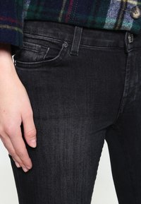 Czarne dżinsy z denimu o dopasowanym kroju, wyposażone w standardowe kieszenie i metalowy guzik w pasie. Gładka tekstura i subtelny połysk.