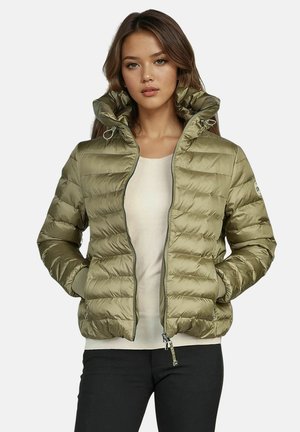 Frau trägt eine olivgrüne Steppjacke mit hohem Kragen, beigen Oberteil und schwarze Hose, steht mit den Händen in den Taschen.