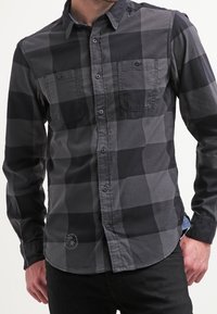Schwarz-grau karierte langärmelige Hemd, aus Baumwollmaterial, Knopfverschluss, zwei Brusttaschen, abgerundeter Saum, mit dezentem Logodetail am unteren Rand.