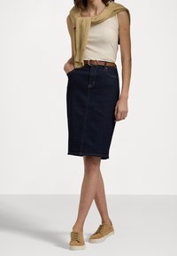 Jupe crayon en denim bleu foncé avec des surpiqûres orange, associée à un débardeur léger et à des baskets beiges à lacets. Accessoirisée avec un pull beige.