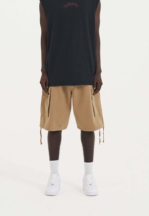Débardeur noir sans manches avec texte rouge, shorts cargo beiges avec poches, et baskets blanches. Tissu texturé et cordons de serrage ajustables présents.