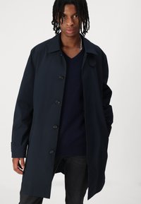 Marineblauwe regenjas met een knoopsluiting aan de voorkant, een iets oversized pasvorm en een brede kraag. Gemaakt van een gladde, waterafstotende stof.