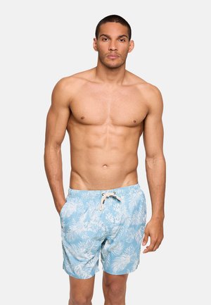 Hombre musculoso de pie con las manos en los bolsillos, vistiendo pantalones cortos de baño azul claro con estampado blanco de hojas tropicales y piña.
