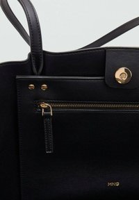 Bolso tote de cuero negro con textura suave, que presenta un bolsillo con cremallera, acentos de herrajes dorados y un cierre a presión. Logotipo de la marca visible.