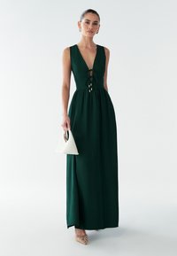 Maxi-jurk in donkergroen met een diepe V-hals en vetersluiting aan de voorkant. Gladde stof, uitlopende rok, gecombineerd met een witte handtas en sierlijke schoenen.