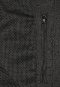 Juodas sportinis audinys su tekstūruota tinkline dalimi ir užtrauktuku uždaromu kišenė. Savybės apima lygias ir kvėpuojančias medžiagas su sluoksniuota konstrukcija.