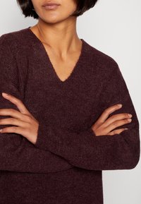 Burgundyröd stickad tröja med V-hals, ribbad textur och långa ärmar. Tyget ser mjukt och mysigt ut, lämpligt för vardagligt bruk.