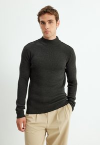 Dunkelgrauer gerippter Pullover mit hohem Kragen und langen Ärmeln, aus einem strukturierten Strickstoff gefertigt. Kombiniert mit beigen Hosen.