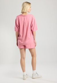 Camisa de manga corta de color rosa claro y pantalones cortos a juego, hechos de tela suave. Presenta un corte relajado, dobladillo redondeado y sutiles detalles de costura.
