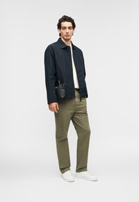 Ελαφρύ μπουφάν ναυτικού χρώματος πάνω από κρεμ πουλόβερ, ελαιδί παντελόνια και λευκά αθλητικά παπούτσια. Μικρή μαύρη τσάντα χιαστί. Το μοντέλο στέκεται μπροστά σε ουδέτερο φόντο.