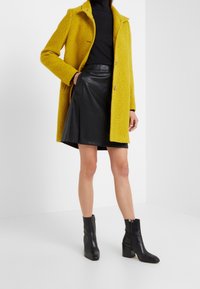 Sinapinkeltainen teksturoitu takki lovikauluksella, kaksi nappia, yhdistetty mustaan nahkahameeseen ja mustiin nilkkureihin.