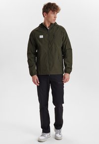 Ljusgrön lättviktsjacka med huva, zip-fickor och elastiska ärmslut. Bärs över svarta byxor och vita sneakers.