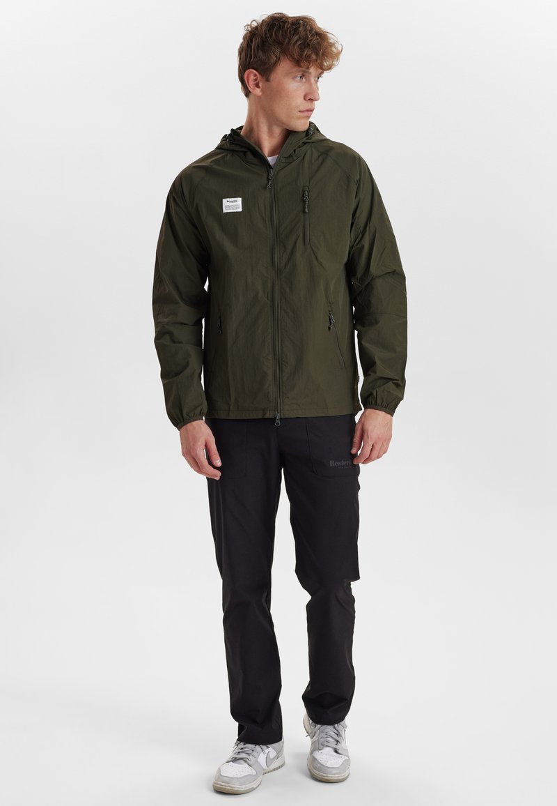 Ljusgrön lättviktsjacka med huva, zip-fickor och elastiska ärmslut. Bärs över svarta byxor och vita sneakers.