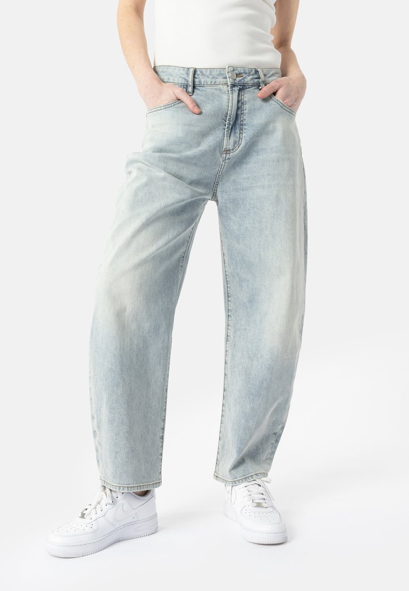 Personne portant un jean bleu clair à coupe ample et des baskets blanches, les mains reposant dans les poches avant, debout sur un fond blanc.