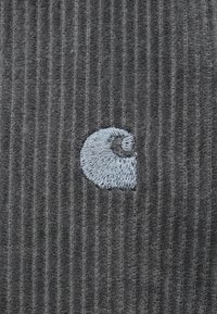 Szürke kordbársony anyag, függőleges bordázott textúrával. Jellemzi egy világoskék hímzett logó, amely a szövet jobb középtáján található.