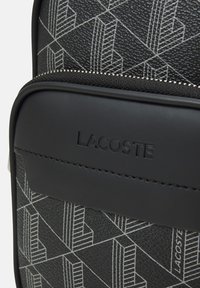 Black textured material backpack with white geometric pattern, featuring a black accent flap and embossed "LACOSTE" logo on the front.