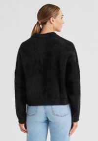 Schwarze, samtige Jacke mit cropped Schnitt, ausgestattet mit einem Kragen und Knopfakzenten an den Ärmeln. Getragen über hellen Jeans.