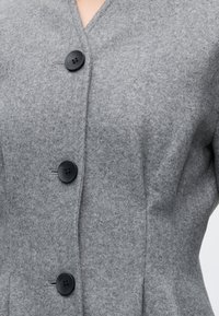 Manteau en laine gris avec trois gros boutons noirs boutonnés sur le devant sur un fond uni.