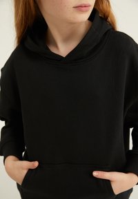 Melna kapuce ar priekšējo kabatu, mīksta auduma tekstūra un brīva piegriezuma ar dziļu izgriezumu. Nav redzamu rakstu vai akcentu.