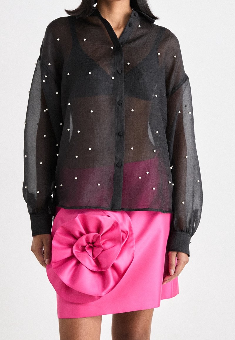 Femme portant un chemisier noir transparent avec de petites perles blanches et une jupe rose vif avec un grand détail de fleur à volants.
