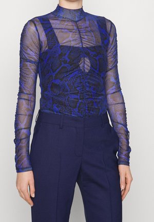 Femme portant un haut transparent à manches longues bleu et noir à motif serpent, par-dessus un caraco noir, associé à un pantalon taille haute bleu foncé.