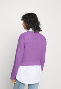 Fioletowy, dziergany sweter krótki z fakturą prążkowaną, okrągłym dekoltem i długimi rękawami, noszony na białej koszuli i zestawiony z jasnoniebieskimi dżinsami.