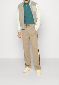 Veste universitaire grise et blanche avec une lettre "G", polo teal et pantalon à carreaux marron avec cordon de serrage et bandes latérales. Baskets jaunes.