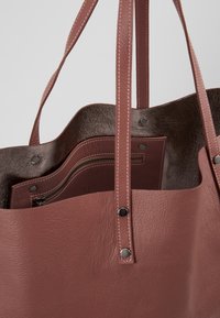 Nahkainen olkalaukku pölyisen vaaleanpunaisena, jossa sileä pinta, kaksi pitkää kantokahvaa ja ruskea sisus, jossa on vetoketjullinen tasku ja napilliset yksityiskohdat.