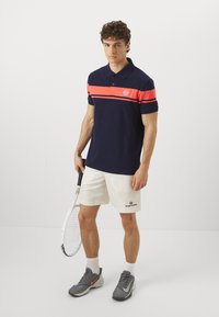 Námořní pánské polo tričko s horizontálním oranžovým pruhem, krátké rukávy a límec. Kombinované s béžovými šortkami a šedými sportovními botami. Drží tenisovou raketu.