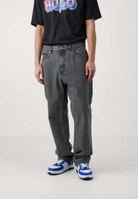 Svart grafisk t-shirt med färggående "HUGO" tryck, i kombination med grå raka jeans. Skorna består av blå och vita sneakers.