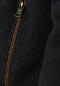 Dunkelblauer Strickstoff mit einem strukturierten Muster, ausgestattet mit einem braunen Akzent-Reißverschluss. Der Stoff wirkt weich und leicht dehnbar.