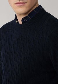 Maglione blu navy a maglia intrecciata con colletto a coste, indossato sopra una camicia a quadri scuri. Presenta motivi testurizzati e una forma aderente.