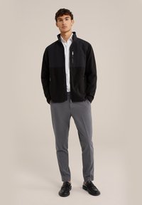 Svart och grå zip-up-jacka med ståkrage, i kombination med gråbyxor och svarta loafers. Har en zip-ficka på vänster bröst.