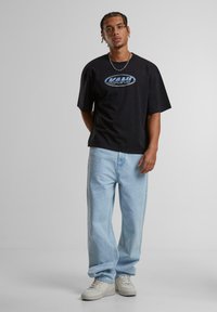Black short-sleeve t-shirt with blue "KANI" logo, paired with light blue relaxed-fit jeans and white sneakers, standing against a plain background.