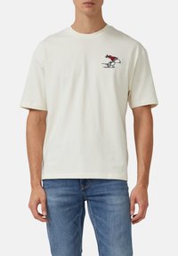 Mann trägt ein weißes kurzärmliges T-Shirt mit einem kleinen gestickten laufenden Snoopy in einem roten Hut auf der linken Brust, kombiniert mit blauen Jeans.