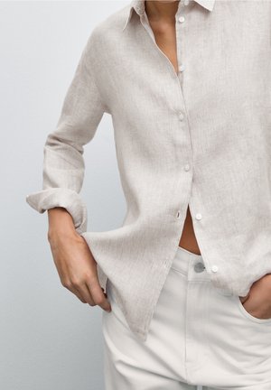 Lichtbeige linnen shirt met een knoopsluiting aan de voorkant, lichtjes textuur, relaxed fit en opgerolde mouwen. Gecombineerd met witte broek.