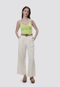 Top senza maniche verde neon con bordi in pizzo e bottoni, abbinato a pantaloni larghi bianchi a vita alta e una cintura intrecciata marrone con scarpe con plateau.
