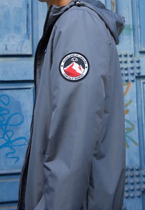 Osoba w szarej kurtce z kapturem z okrągłą naszywką narciarską na górnej części ramienia, stojąca na tle niebieskich drzwi pokrytych graffiti.