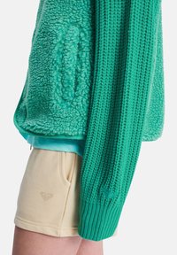 Zielona kurtka z polaru z teksturowanym, puszystym materiałem i ściągaczowymi rękawami; noszona na jasnoniebieskim topie i beżowych szortach.